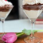 A mousse cup featuring layers of chocolate velvet mousse and mascarpone cream is topped with whipped cream and a raspberry, beside a pink tulip on a wooden surface. Another dessert glass appears softly blurred in the background.