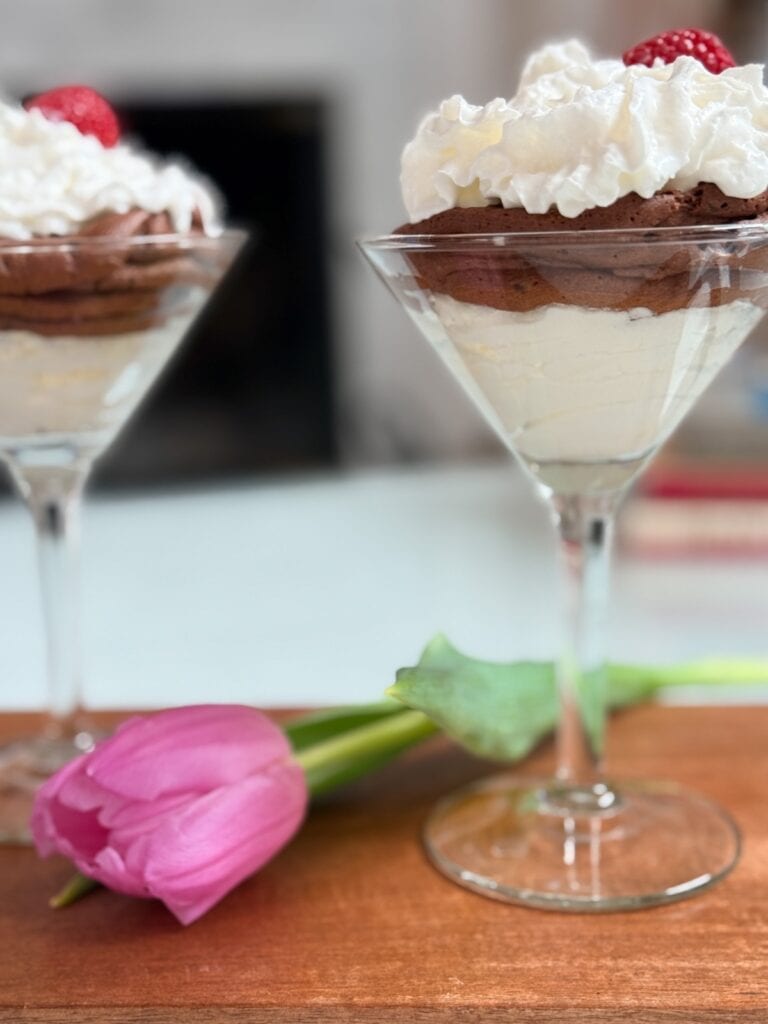 A mousse cup featuring layers of chocolate velvet mousse and mascarpone cream is topped with whipped cream and a raspberry, beside a pink tulip on a wooden surface. Another dessert glass appears softly blurred in the background.