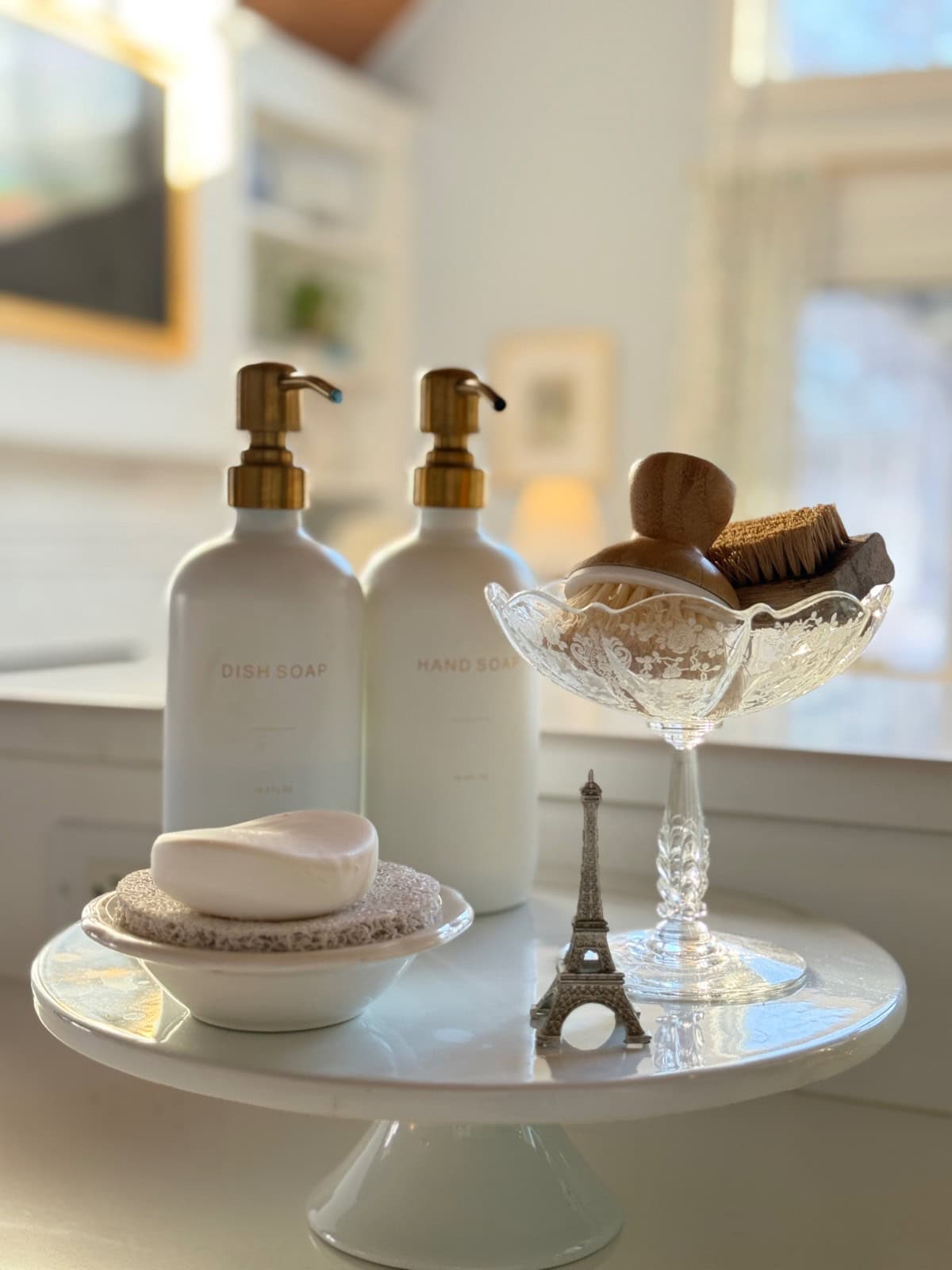A kitchen counter displays the latest bottles of dish soap and hand soap, a soap bar on a dish, wooden cleaning brushes in a glass bowl, and a small Eiffel Tower figurine, all arranged on a white pedestal stand. Sunlight streams in softly.