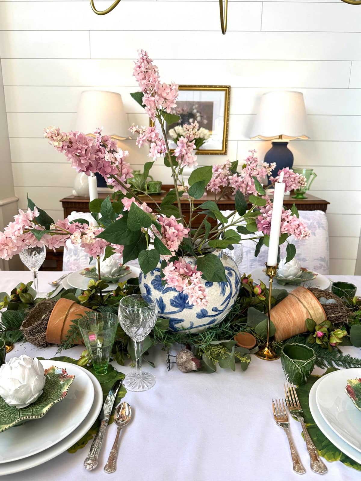 A spring-themed table setting features a blue and white vase with pink lilac flowers, surrounded by green foliage, glassware, candles, shopping bags, garden tools, and potted plants on a white tablecloth. Lamps and a framed photo are in the background.