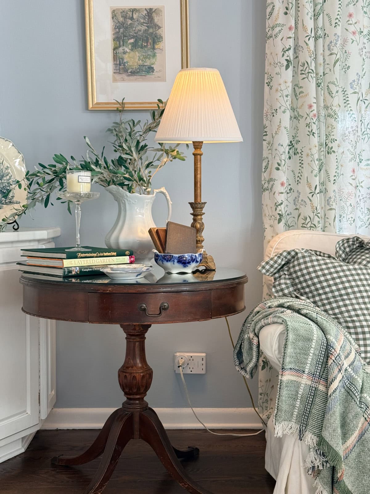 A round wooden side table with books, a lamp, a pitcher with greenery, and a bowl sits next to a white sofa draped with the latest green checked blanket and floral curtains, creating a cozy, vintage-inspired living space.