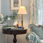 A cozy living room corner perfect for downsizing to a smaller home, featuring a round wooden table with a lamp, books, and greenery. A white sofa with plaid pillows and floral curtains creates an inviting, space-saving retreat.