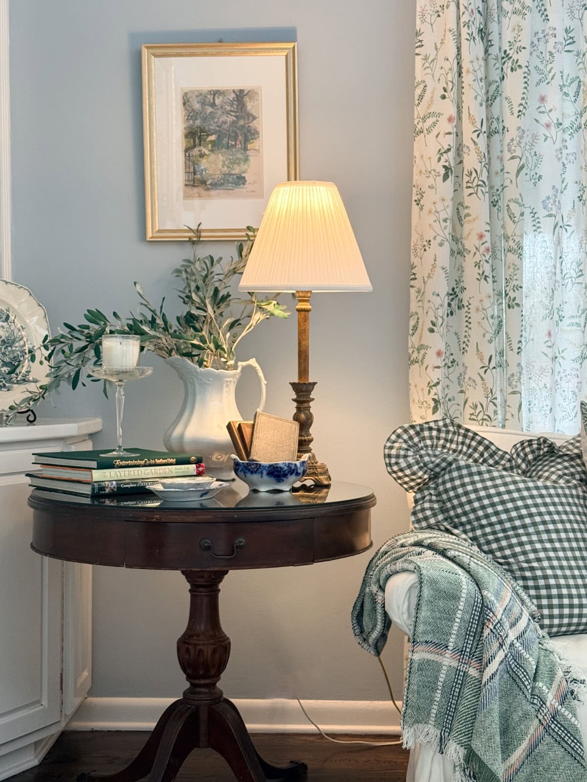 A cozy living room corner perfect for downsizing to a smaller home, featuring a round wooden table with a lamp, books, and greenery. A white sofa with plaid pillows and floral curtains creates an inviting, space-saving retreat.