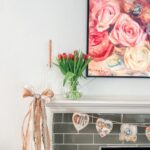 An elegant Valentine’s Day decor: a mantel adorned with a vase of red tulips, a pink candle, a large ribbon, and a heart-shaped garland, all beneath a floral painting of pink and white roses.
