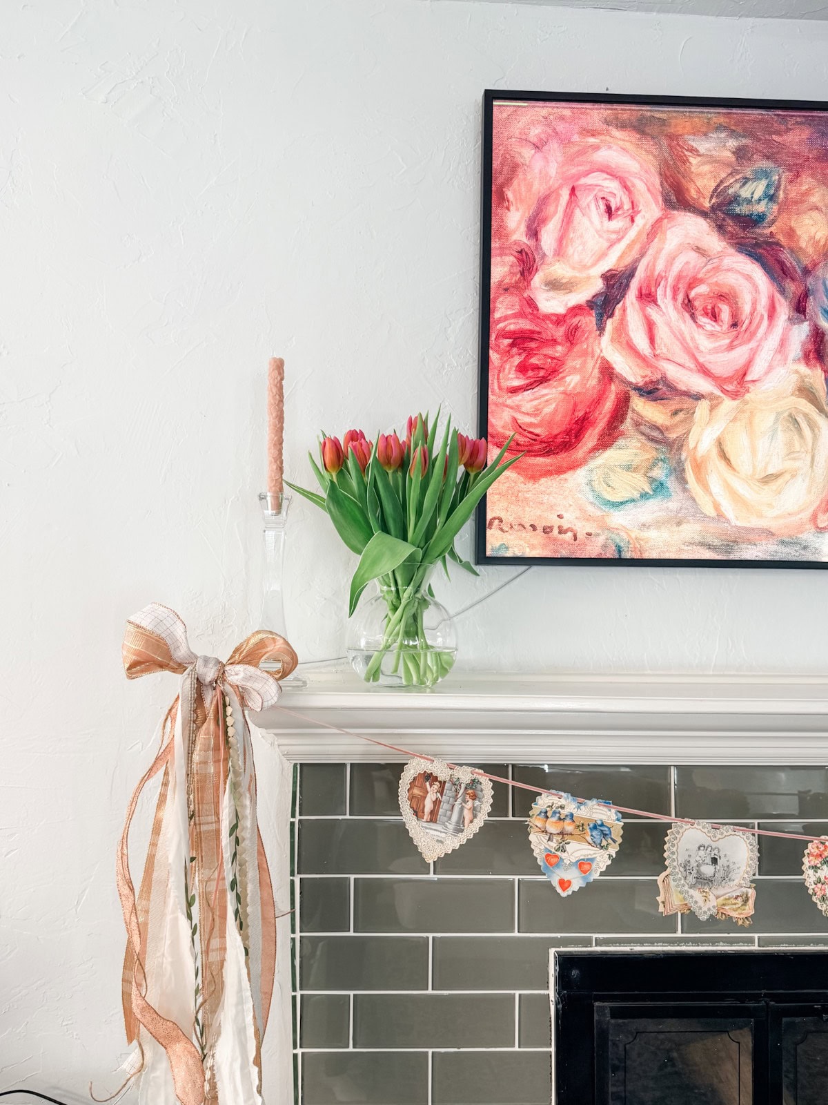 An elegant Valentine’s Day decor: a mantel adorned with a vase of red tulips, a pink candle, a large ribbon, and a heart-shaped garland, all beneath a floral painting of pink and white roses.