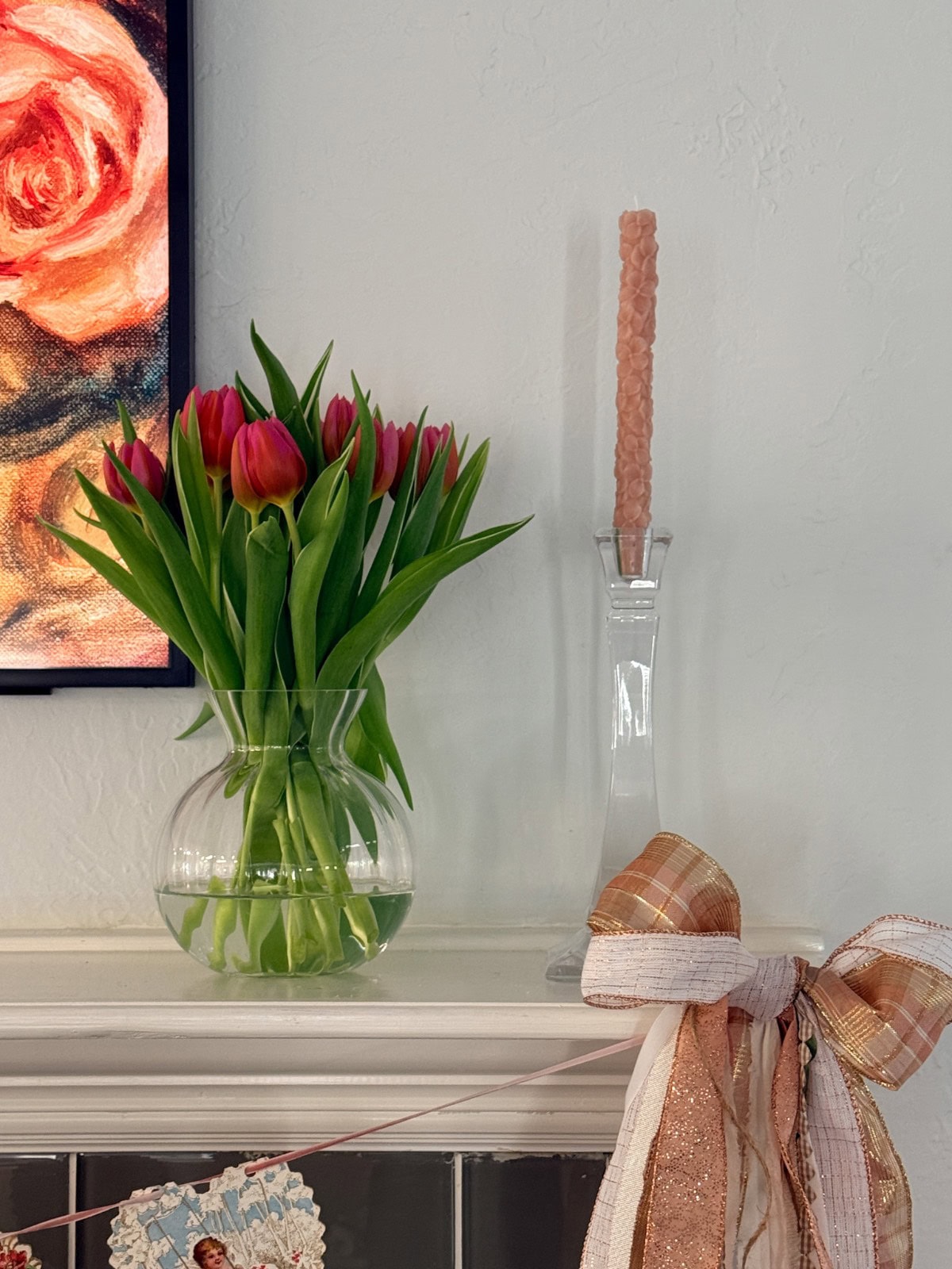 A clear vase of pink tulips and a pink textured candle in a glass holder are displayed on a white mantel, evoking memories of shopping with friends. A plaid ribbon and part of a floral painting are also visible.