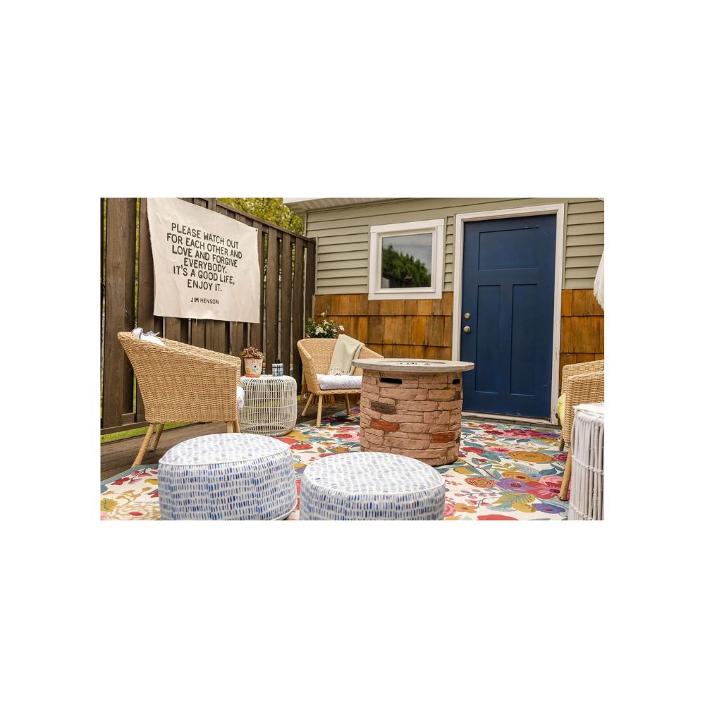 Cozy outdoor patio with wicker chairs, patterned ottomans, a round stone fire pit, colorful rug, and a blue door. Perfect for your next House Tour, it features a cloth sign with a quote on the fence for a welcoming touch.
