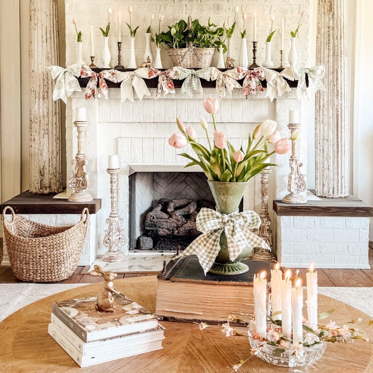 A cozy living room featured in this Home Tour boasts a white brick fireplace adorned with candles, greenery, and floral garlands. Pink tulips and stacked books sit on a round wooden table, with lit candles and a bunny figurine nearby.