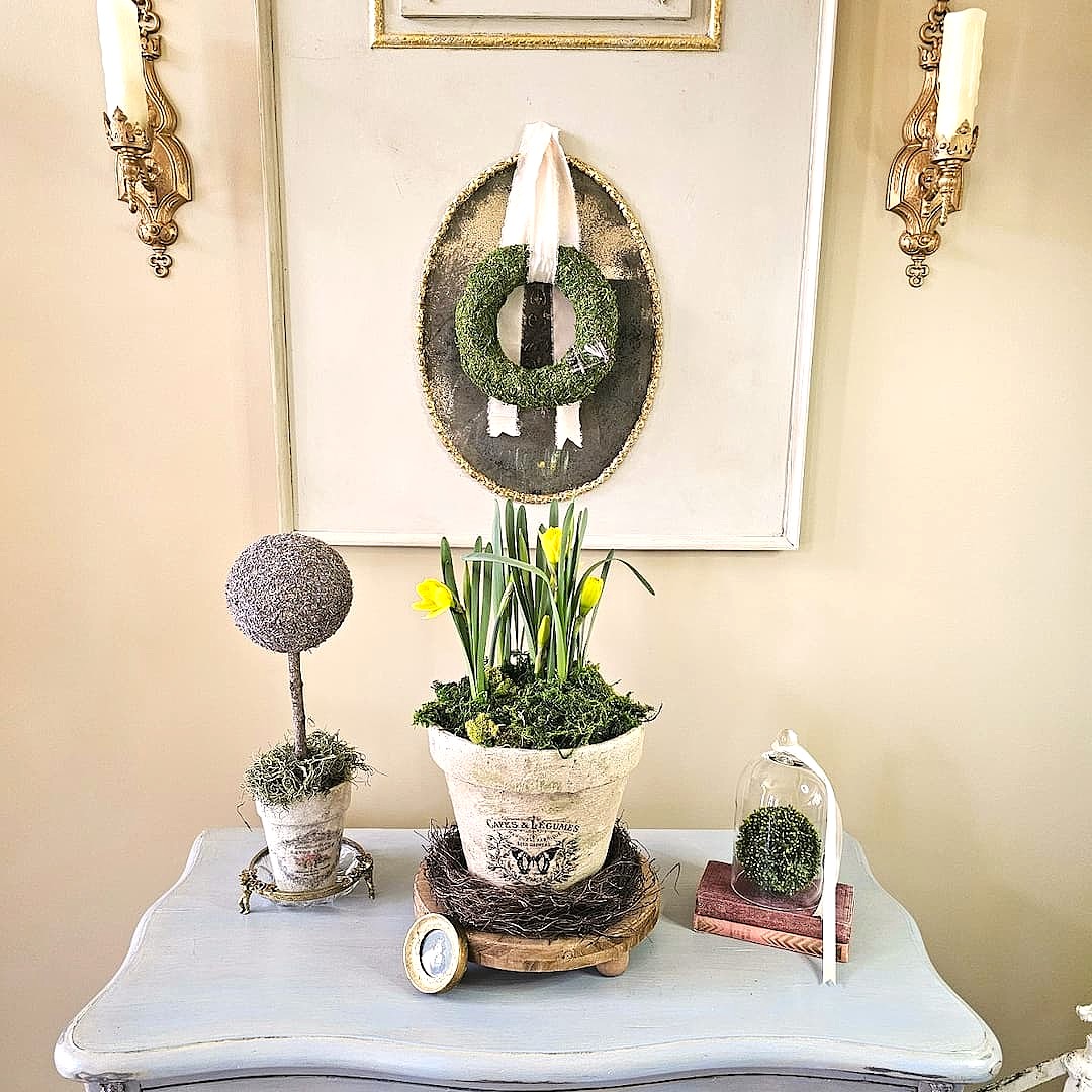 A light blue table holds a potted plant with yellow flowers, a moss topiary, a small clock, and a glass cloche covering a moss ball; above hangs a green wreath on a white-framed backdrop—perfect for any Home Tour Code inspiration.