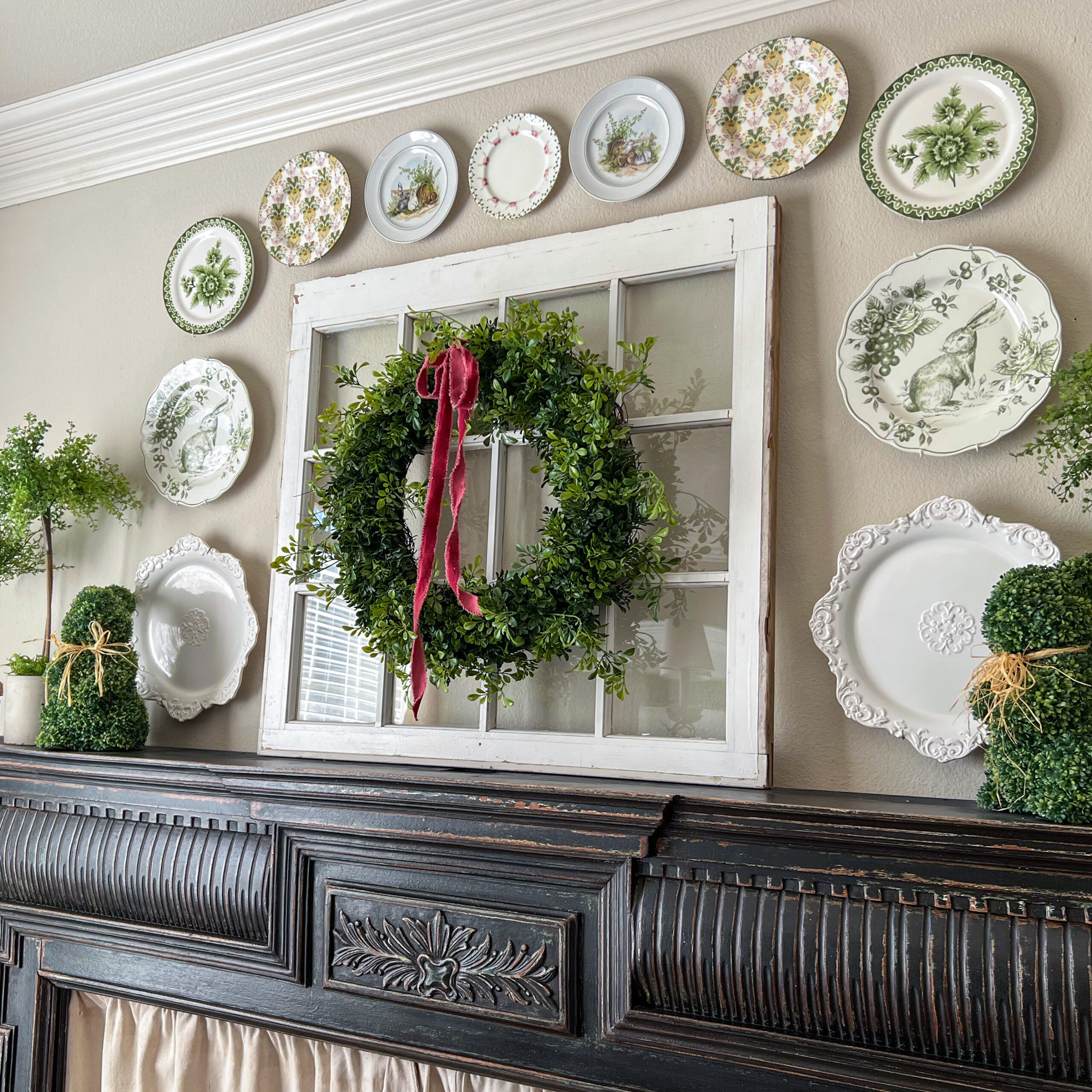 A decorative mantel, perfect for your next Home Tour, features a vintage window with a wreath and red ribbon, flanked by greenery and white plates with floral designs artfully arranged on the wall above.
