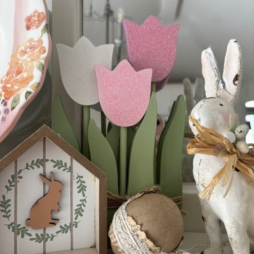 A spring-themed decor scene for your Home Tour, featuring pastel wooden tulips, a white rabbit figurine with a raffia bow, a round ornament with lace, and a small house with a bunny silhouette.