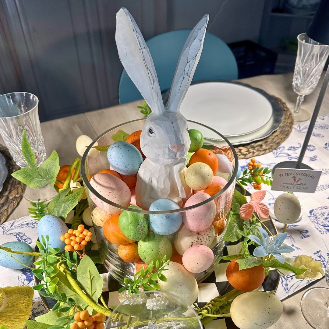 A white ceramic bunny sits in a glass bowl filled with pastel eggs, surrounded by artificial flowers and greenery on a decorated table set for a meal—perfect for your next Home Tour celebration.