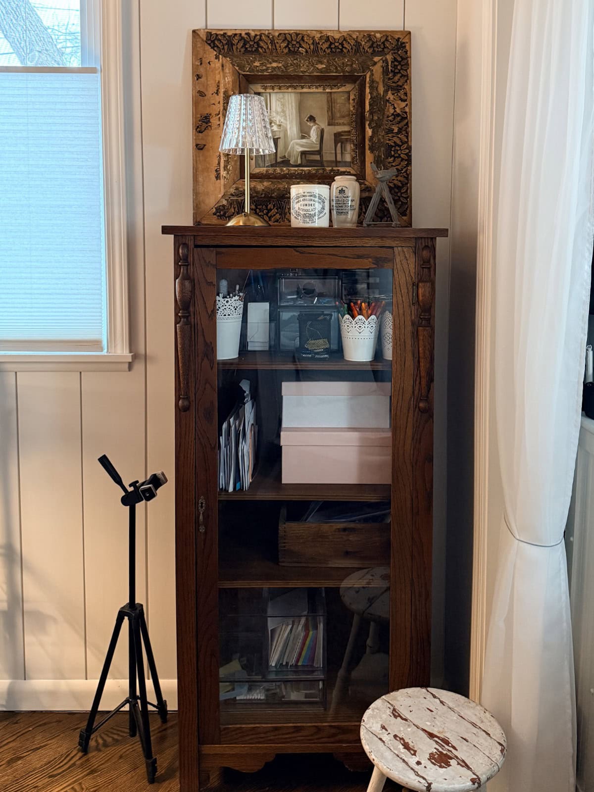A wooden cabinet with glass doors holds books, boxes, and the latest office supplies. On top, a framed photo, small lamp, and decorative items are displayed. A tripod stands nearby; a distressed white stool sits in front of white curtains.