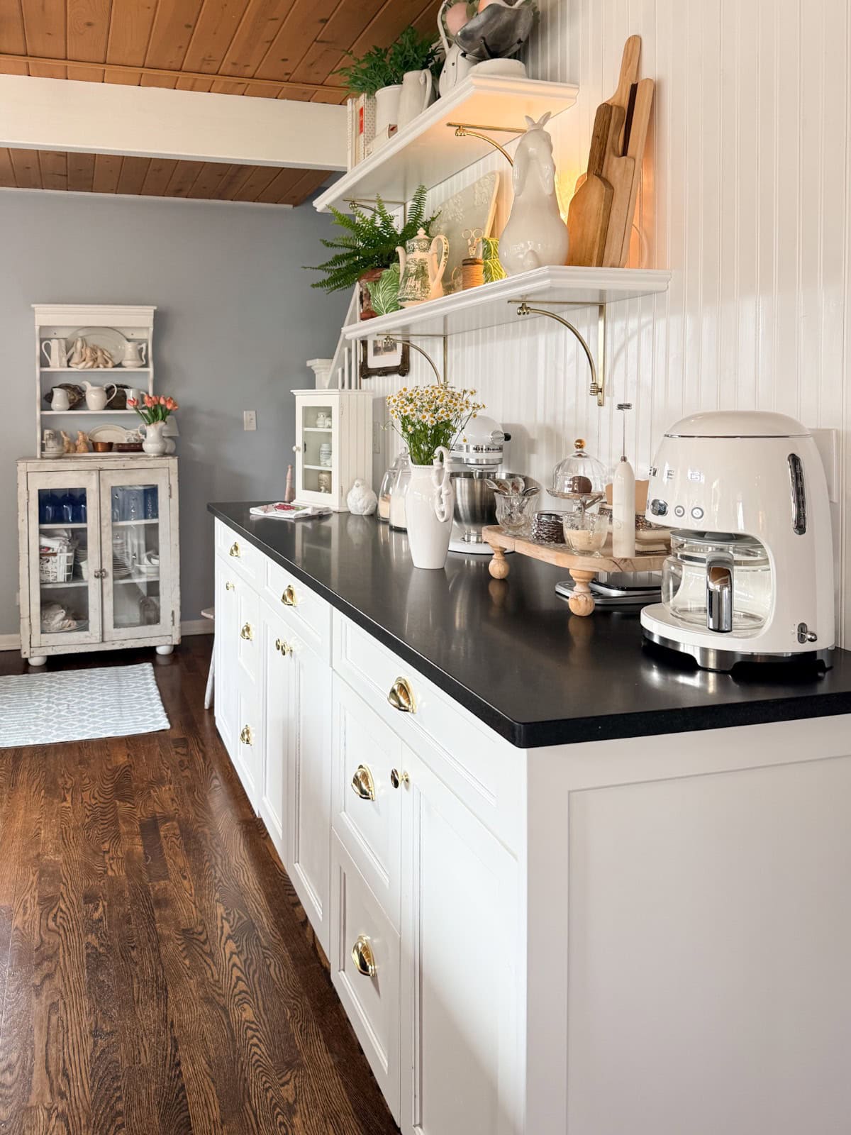 A bright, cozy kitchen with white cabinets, black countertops, open shelves holding dishes and plants, a white coffee machine, and wooden accents. The dark wood floor adds warmth—perfect for a Spring Home Tour or a special Kitchen Reveal.