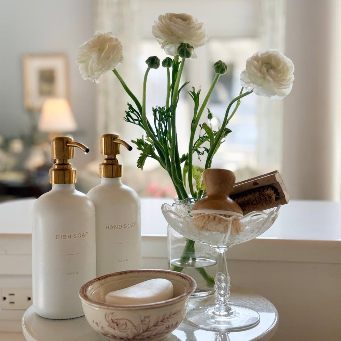 A neatly arranged countertop with bottles of dish soap and hand soap, a bowl with a bar of soap, a glass dish holding brushes, and a vase of white ranunculus flowers—this space is pure Home Tour Code inspiration.