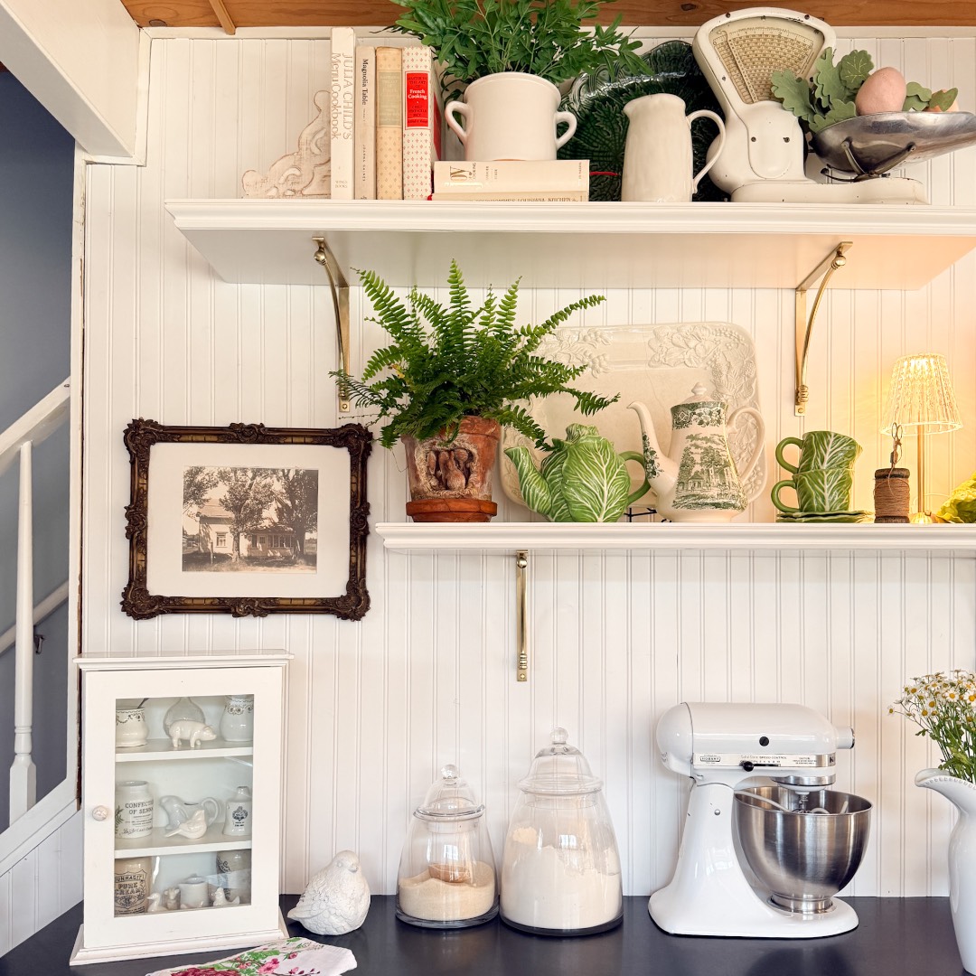 A cozy, rustic kitchen corner with white shelves displays books, dishes, a potted fern, vintage decor, and jars of flour and sugar—perfect for a Spring Home Tour or inspiring your next charming Peter Rabbit tablescape.