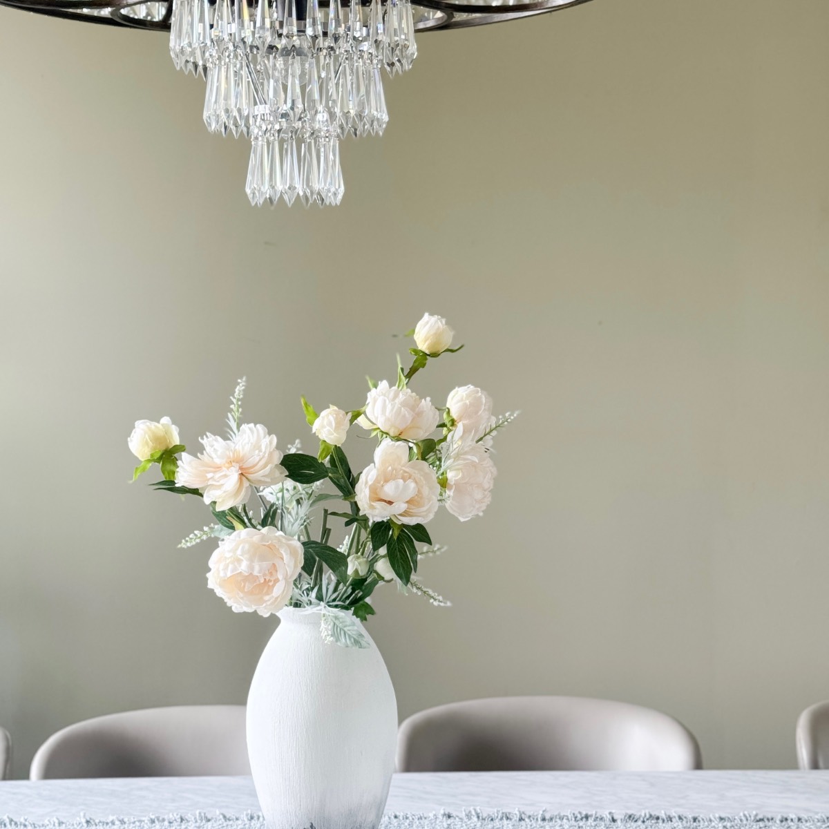A white vase with light pink roses sits on a table with a white cloth, underneath a crystal chandelier. The serene setting offers perfect inspiration for your next Home Tour, set against a plain beige wall and partially visible chairs.