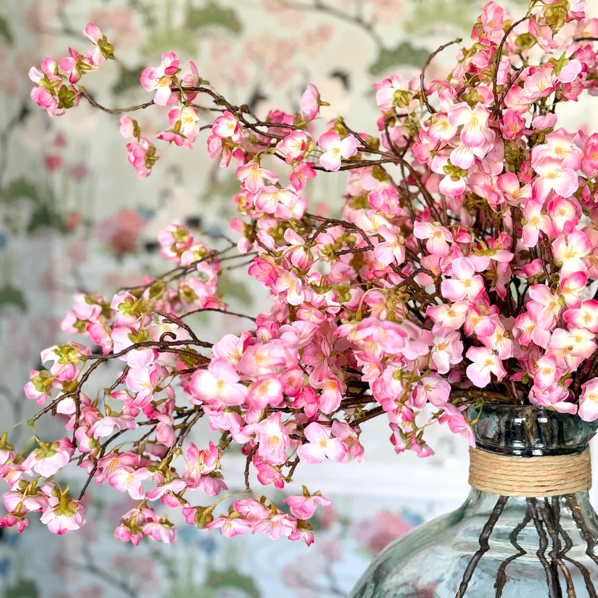 A glass vase filled with branches of vibrant pink cherry blossoms, set against a softly blurred floral-patterned background, creates a stunning scene perfect for any Home Tour.