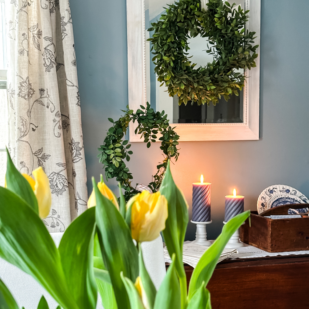 A cozy room with yellow tulips in the foreground, two green wreaths—one hanging on a mirror and one leaning on the wall—and two lit candles on a table beside a wooden box and blue-patterned plates, ready for a Home Tour.