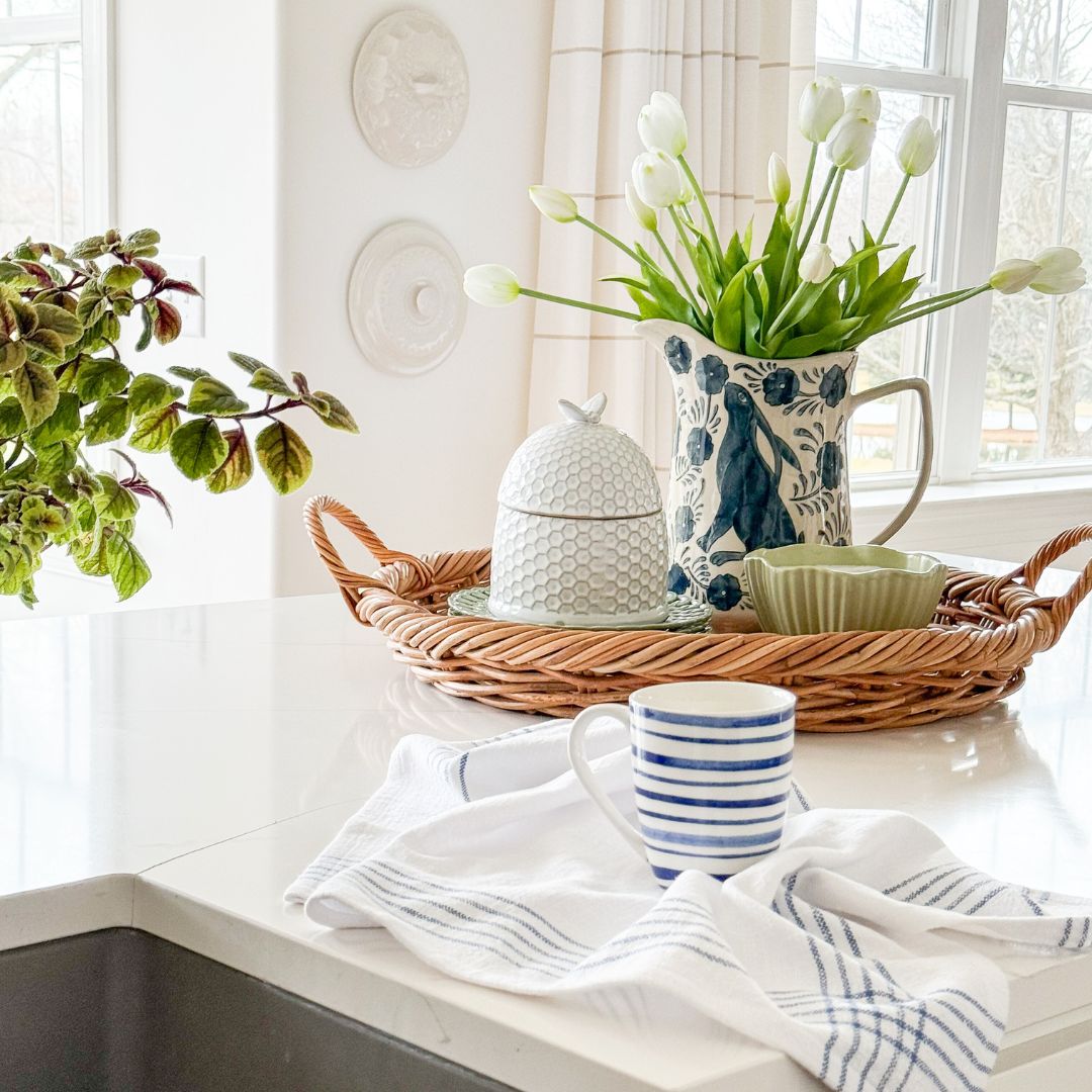 A wicker tray with a floral pitcher of white tulips, a honey jar, and green bowls sits on a white countertop. In front, a striped mug and blue-striped towel are by the sink. Natural light fills the space—perfect for your next Virtual Home Tour.