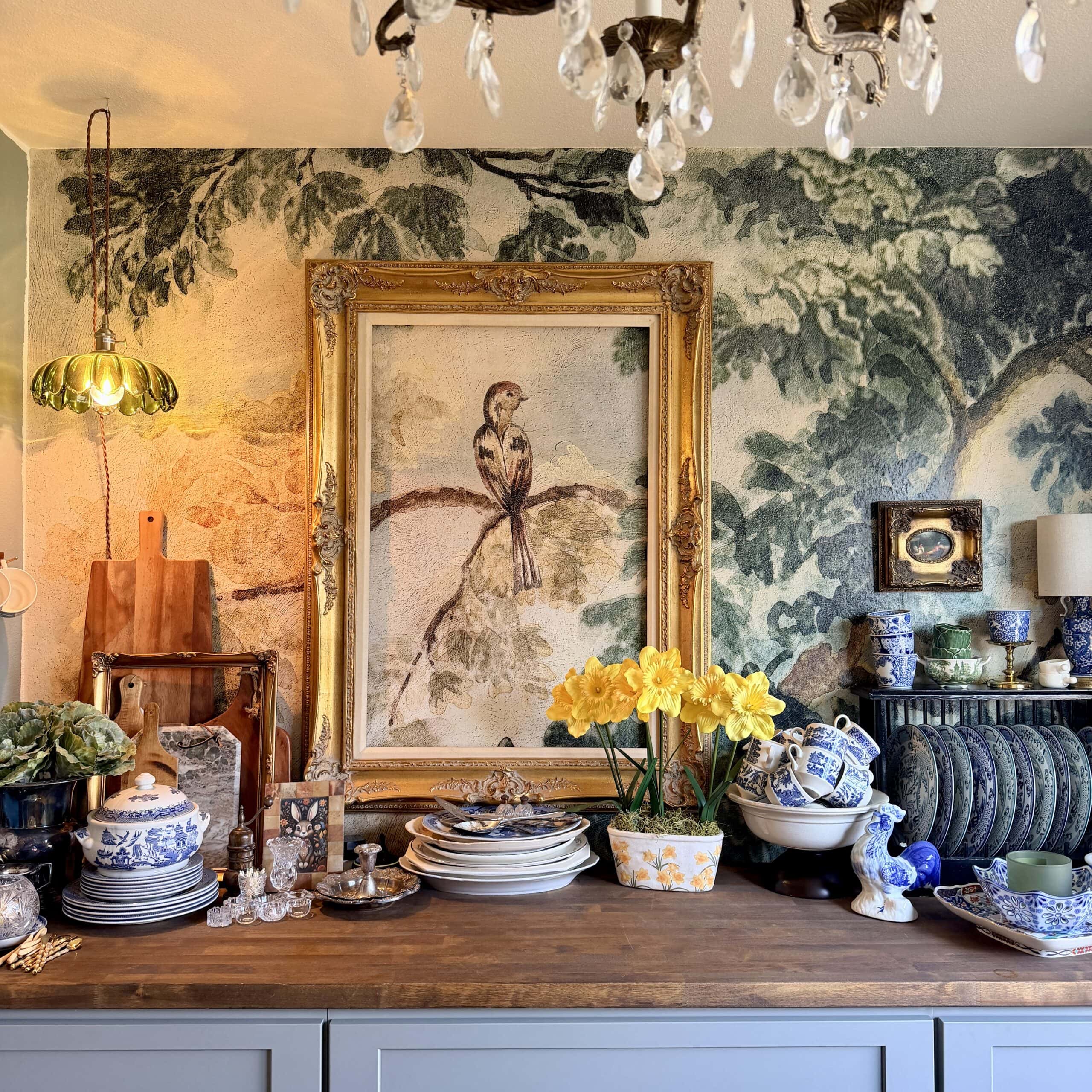 A cozy kitchen scene with a wooden countertop holding blue and white china, crystal glasses, cutlery, yellow daffodils, and decorative items. Perfect for any Home Tour Code, the wall features a large bird painting, leafy wallpaper, and a chandelier overhead.