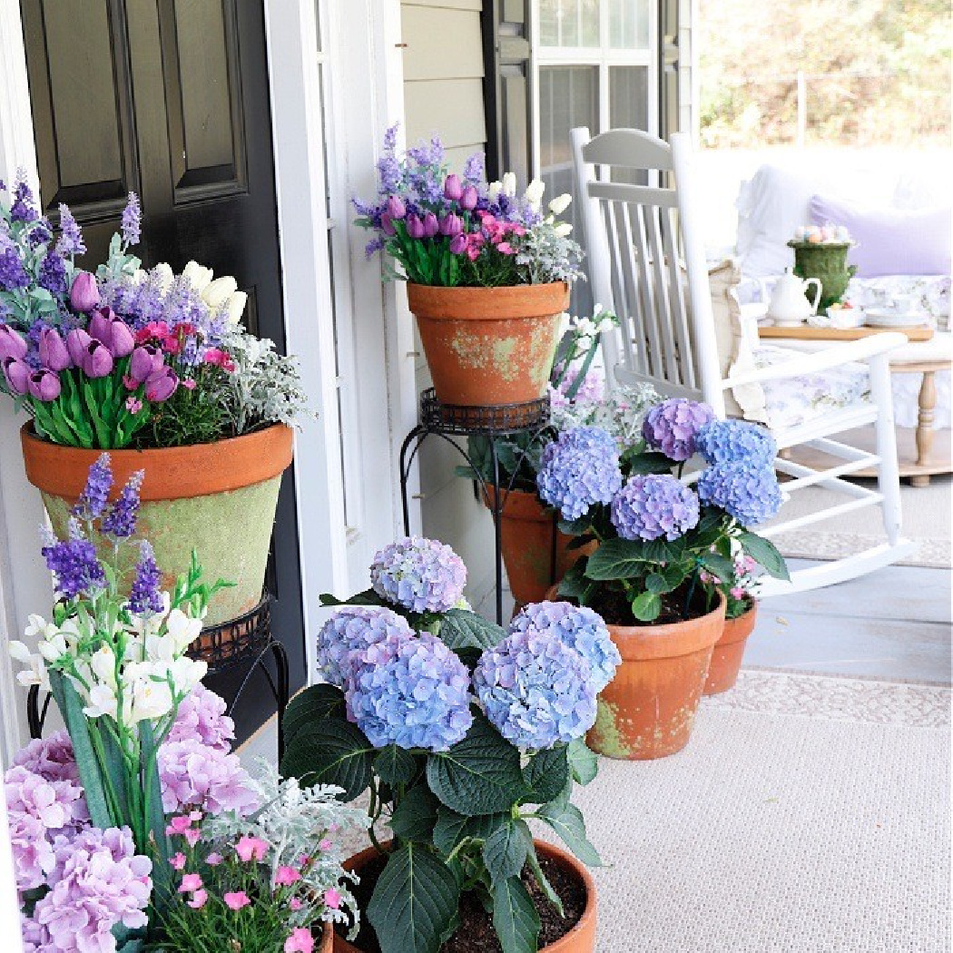 Potted hydrangeas and mixed flowers in terracotta pots decorate a bright porch with a white rocking chair and cushioned bench, setting the scene for a cozy Home Tour in this inviting outdoor seating area.