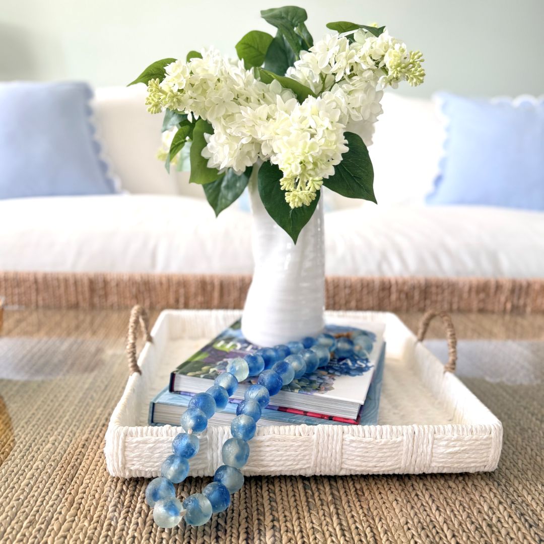 A white vase with white flowers sits on a white tray with books and a blue beaded garland, placed on a woven coffee table. This serene Home Tour setting features a white sofa with light blue pillows in the background.