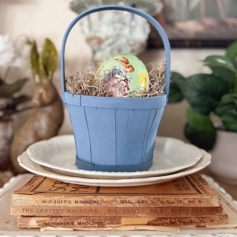 A small blue basket with an Easter egg decorated with a bunny and flowers sits on a white plate, atop vintage magazines—an inviting Home Tour scene, with a plant and brown bunny figurine visible in the blurred background.
