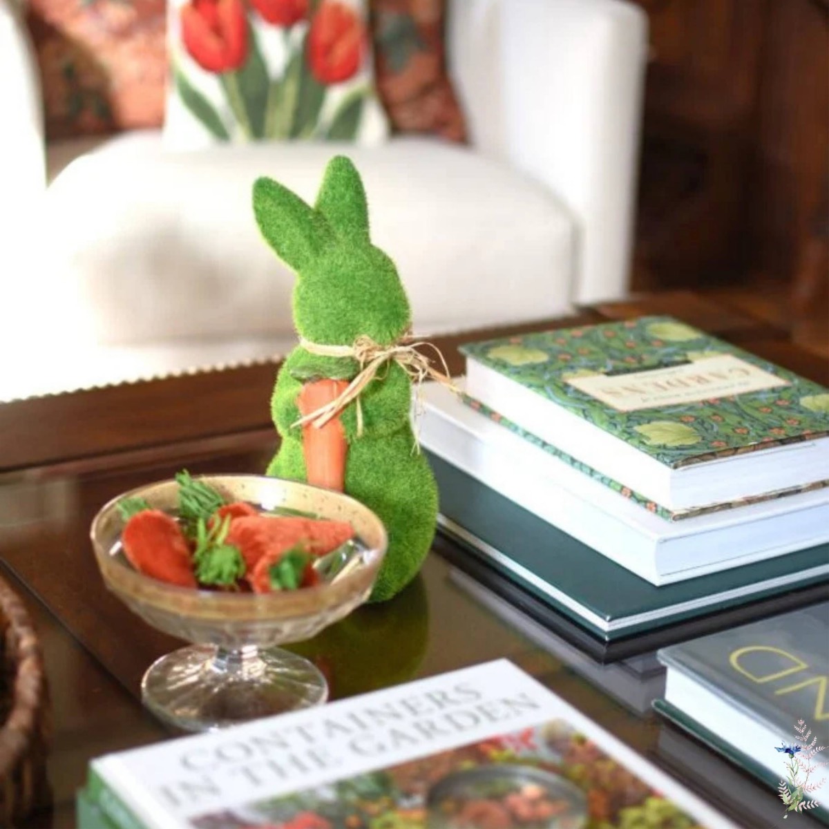 A green, grass-like bunny figurine with a twine bow and carrot sits on a coffee table near books and strawberries—perfect for styling your space or sharing in your next virtual home tour. A chair with a tulip pillow accents the background.