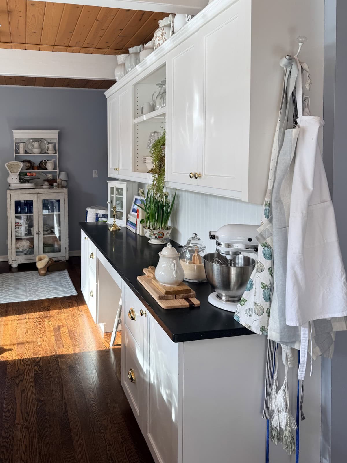 Bright kitchen with white cabinets, black countertop, stand mixer, and teapot. Sunlight streams in, highlighting wooden floors—perfect for a Spring Home Tour. A hutch filled with dishes stands in the background, evoking fresh inspiration for any Kitchen Reveal.