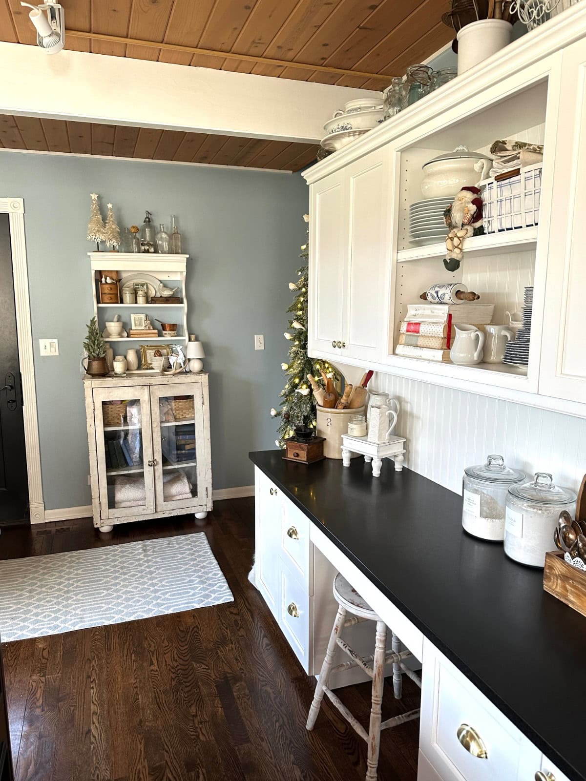 A cozy kitchen reveal with white cabinets, black countertop, and wood floors. Open shelves display dishes and decor. A small Christmas tree and festive decorations add a holiday touch. A white rug lies on the floor.
