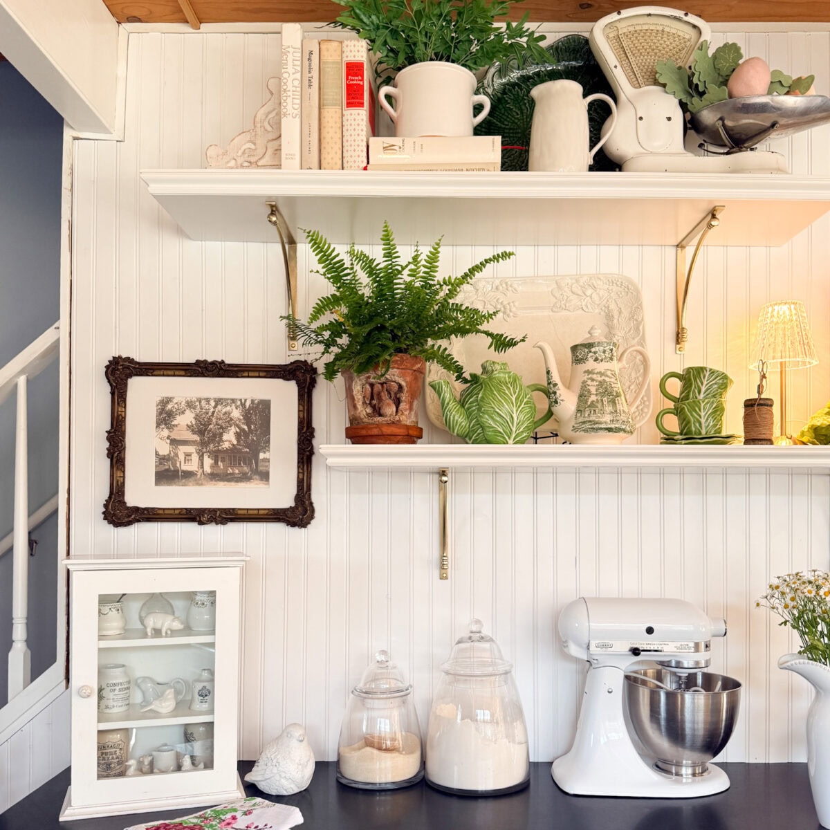 A cozy kitchen corner from our Kitchen Reveal features white shelves with cookbooks, plants, vintage scales, and mugs. Below, a stand mixer and glass jars add charm—perfect inspiration for your Spring Home Tour.