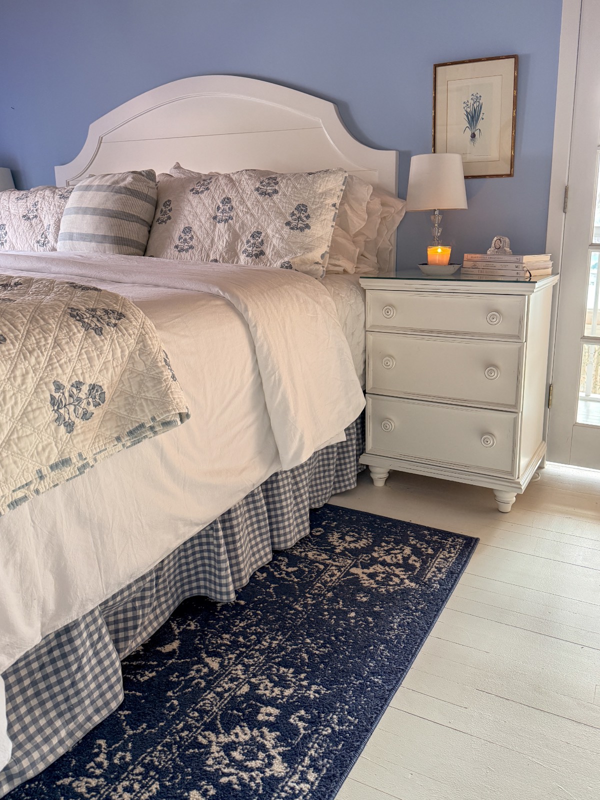 A cozy bedroom inspired by Shopping with Friends, with a white bed featuring floral bedding, a blue and white plaid bed skirt, a blue patterned rug, and a nightstand holding books, a lamp, and a lit candle against a soothing blue wall.