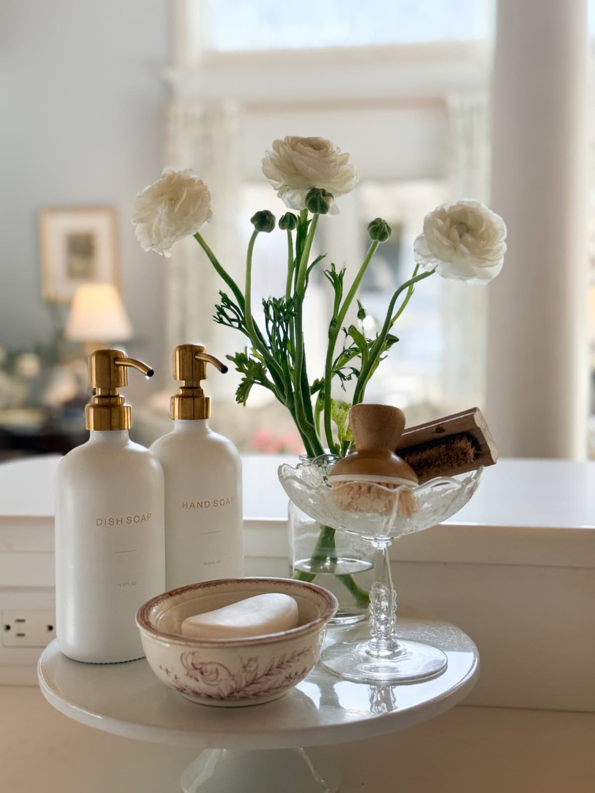 A kitchen counter with white dish soap and hand soap bottles, a bowl with a bar of soap, a glass stand holding a brush and sponge, and a vase with white flowers set the scene for a fresh Spring Home Tour.