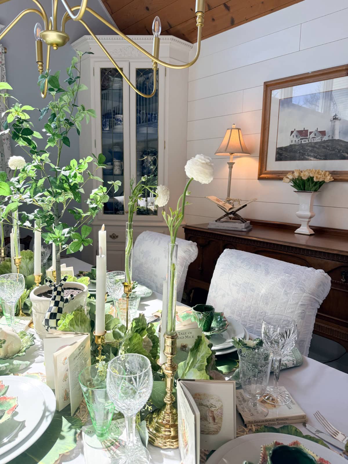 Elegant dining room with a sunlit table set for a meal, featuring vintage Easter table decor—white flowers in vases, green glassware, gold candlesticks, leafy plates, and a lush plant centerpiece. Cozy, bright space with a lamp and framed art in the background.