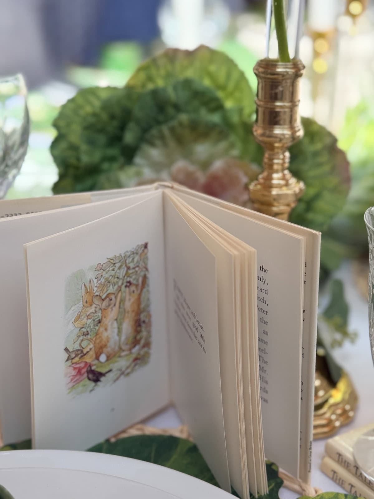 An open illustrated children's book, featuring rabbits on one page, stands on a table decorated with green leaves and glassware, creating a whimsical Peter Rabbit Tablescape and garden-like setting perfect for a Spring Home Tour.
