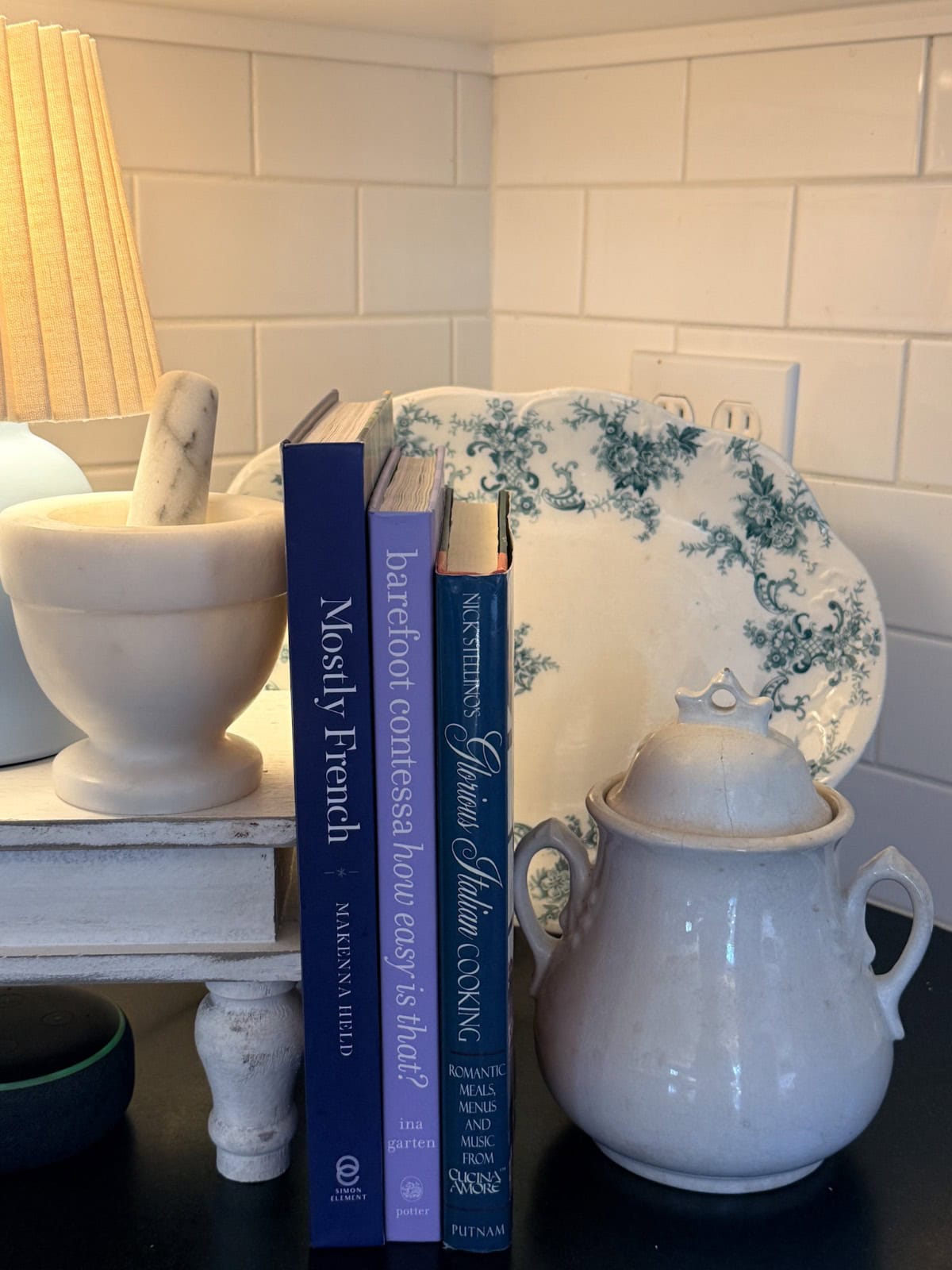 A mortar and pestle, three cookbooks, a decorative plate, and a white ceramic jar with a lid are arranged on a dark kitchen counter—perfect details for any Spring Home Tour—with a white tile backsplash and a lit lamp in the background.
