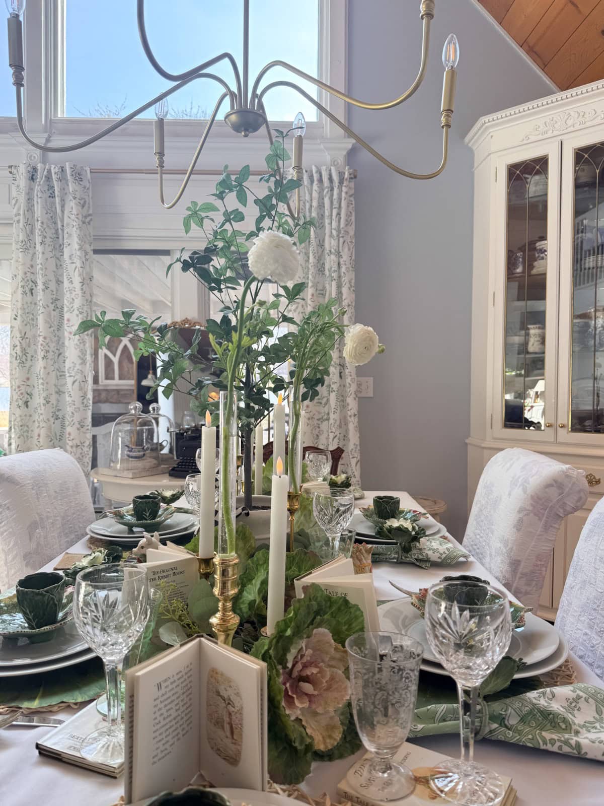 A dining table is elegantly set with green tableware, crystal glasses, white candles, open books, and floral centerpieces—a perfect Peter Rabbit Tablescape. Light streams through large windows with patterned curtains, and a brass chandelier hangs above.