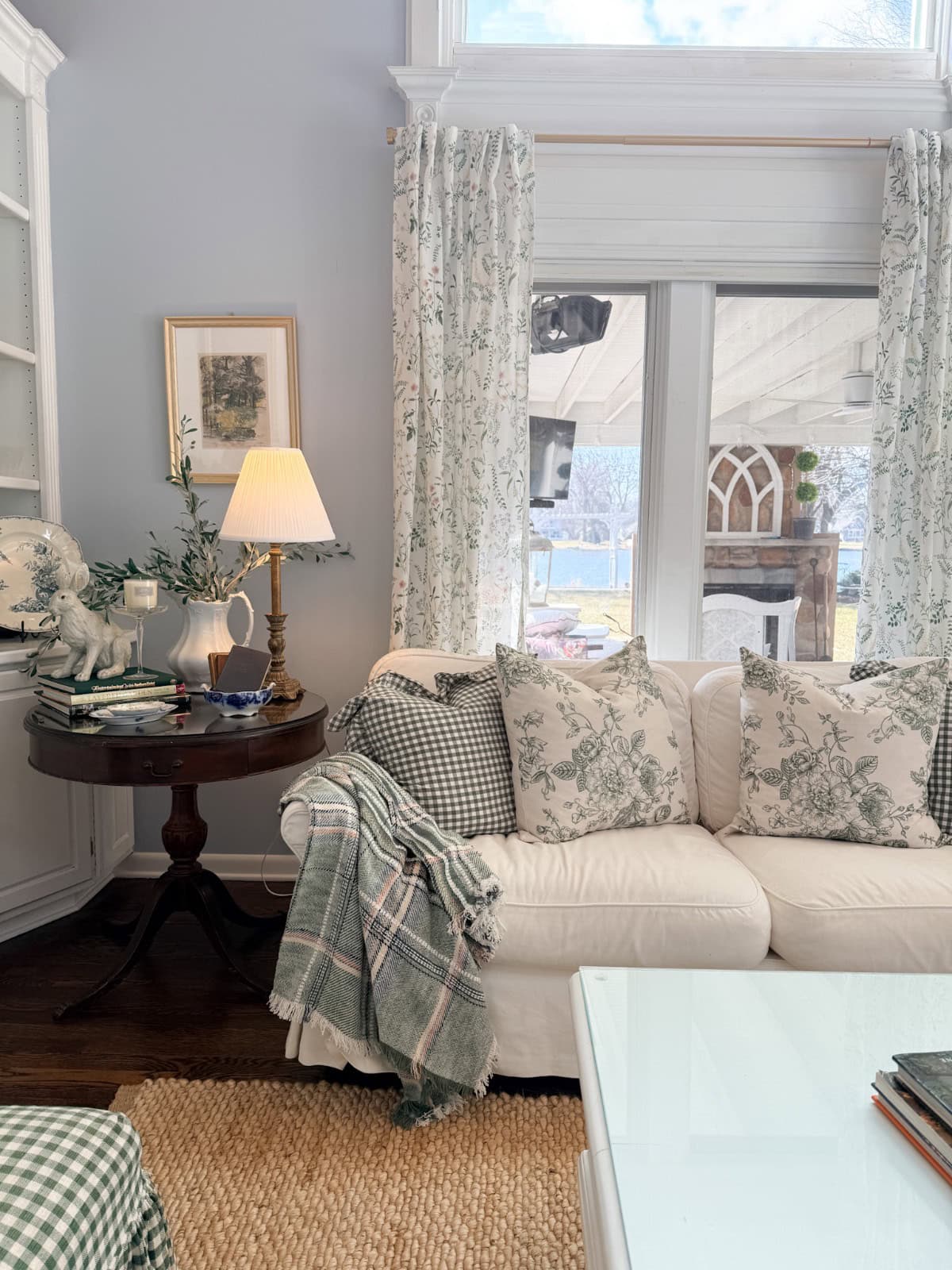 A cozy living room with a white sofa covered in green and white patterned pillows and a plaid throw blanket. A round wooden side table holds a lamp, books, and decorative items—perfect for relaxing after a Spring Home Tour. Large windows with floral curtains let in natural light.