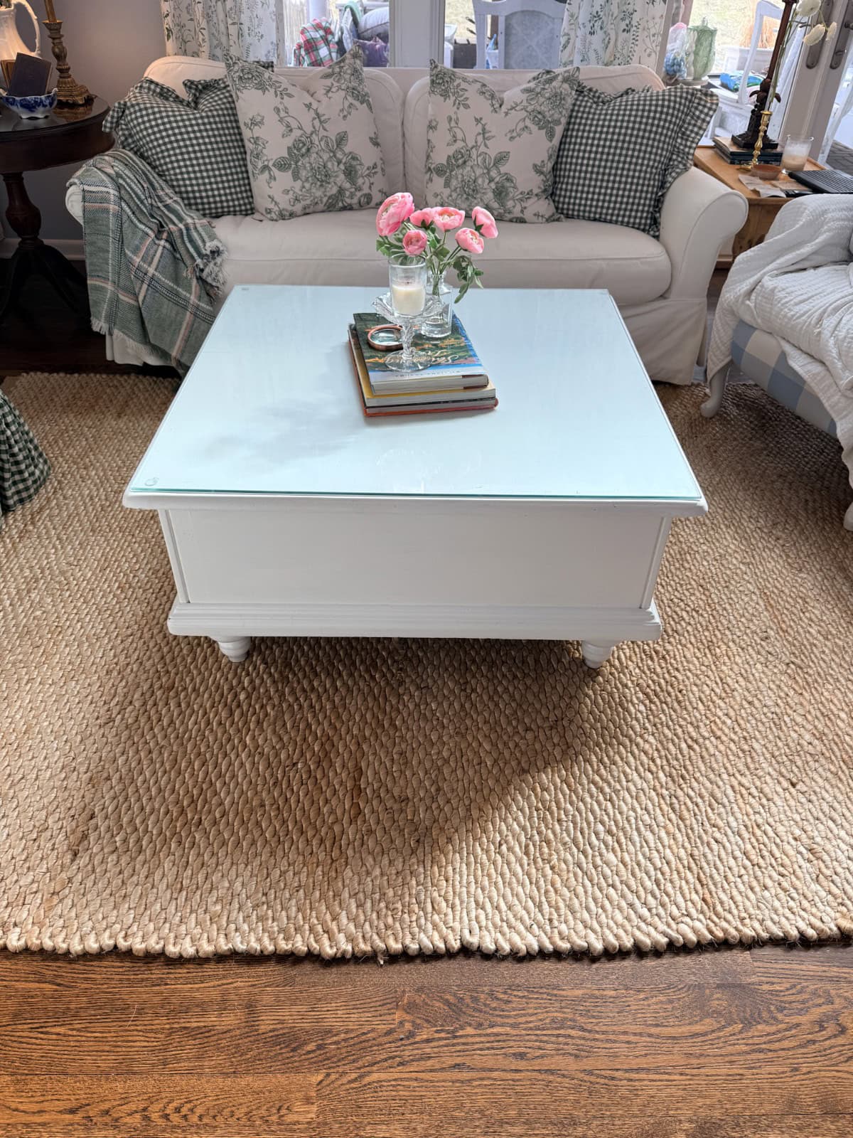 A white coffee table with a glass top sits on a woven rug in front of a white sofa with green and white pillows. Perfect for your Spring Home Tour, the cozy cottage-style room features pink flowers and books, adding fresh charm.