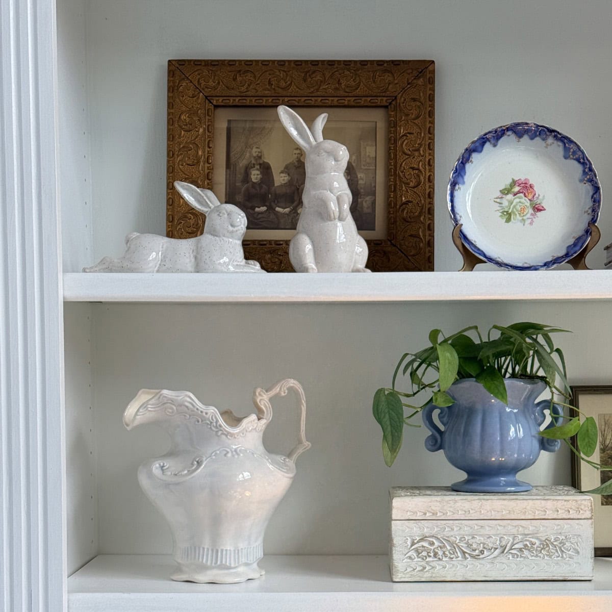 A white shelving unit displays ceramic bunny figurines, a framed vintage photograph, a decorative plate with a floral design, and more—perfect touches for a Spring Home Tour or inspiration for your next Peter Rabbit Tablescape.