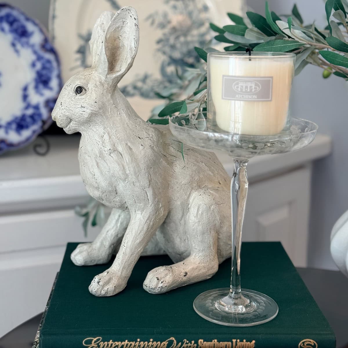 A white rabbit figurine sits on a green book next to a glass candle holder with a cream-colored candle, creating a charming Peter Rabbit Tablescape. Decorative plates and greenery are arranged in the background.