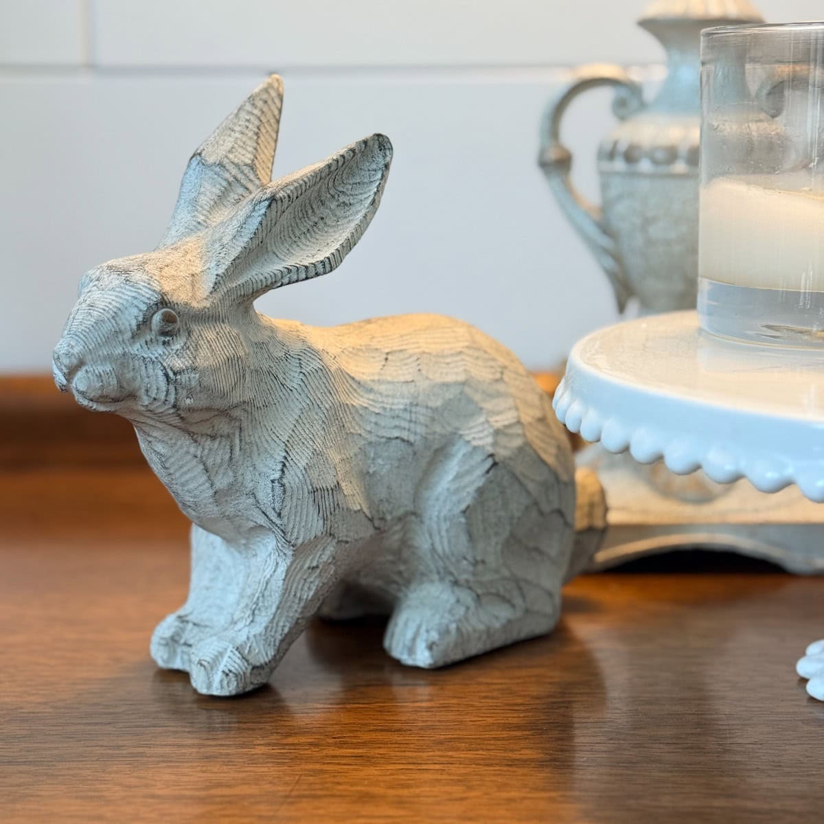 A carved wooden rabbit figurine sits on a wooden surface beside a white cake stand, creating charming Peter Rabbit tablescape vibes, with a glass and a decorative pitcher in the background.