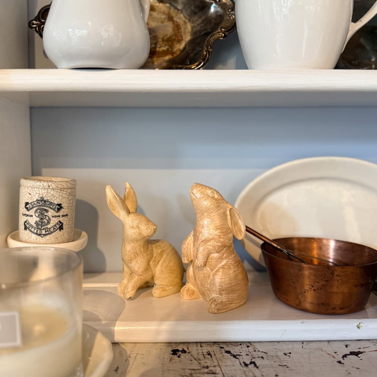 Two carved wooden rabbit figurines sit on a white shelf, surrounded by a copper bowl, a candle, and ceramic dishes—a charming touch for your Peter Rabbit Tablescape or Spring Home Tour.