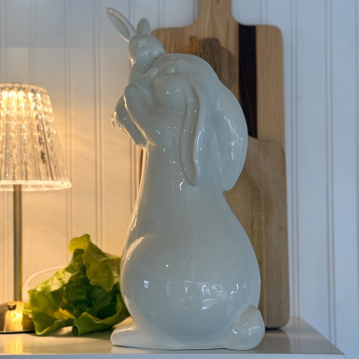 A glossy white ceramic bunny figurine sits upright on a white shelf, with droopy ears and a small bunny on its head—a charming touch for any Spring Home Tour. A lamp, green lettuce, and wooden cutting boards accent the kitchen in the background.
