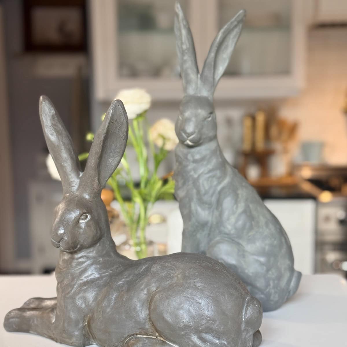 Two gray rabbit sculptures rest on a white surface—one lying down, the other sitting upright—adding charm to a Spring Home Tour. In the background, a kitchen reveal features flowers and blurred cabinets for a fresh seasonal vibe.