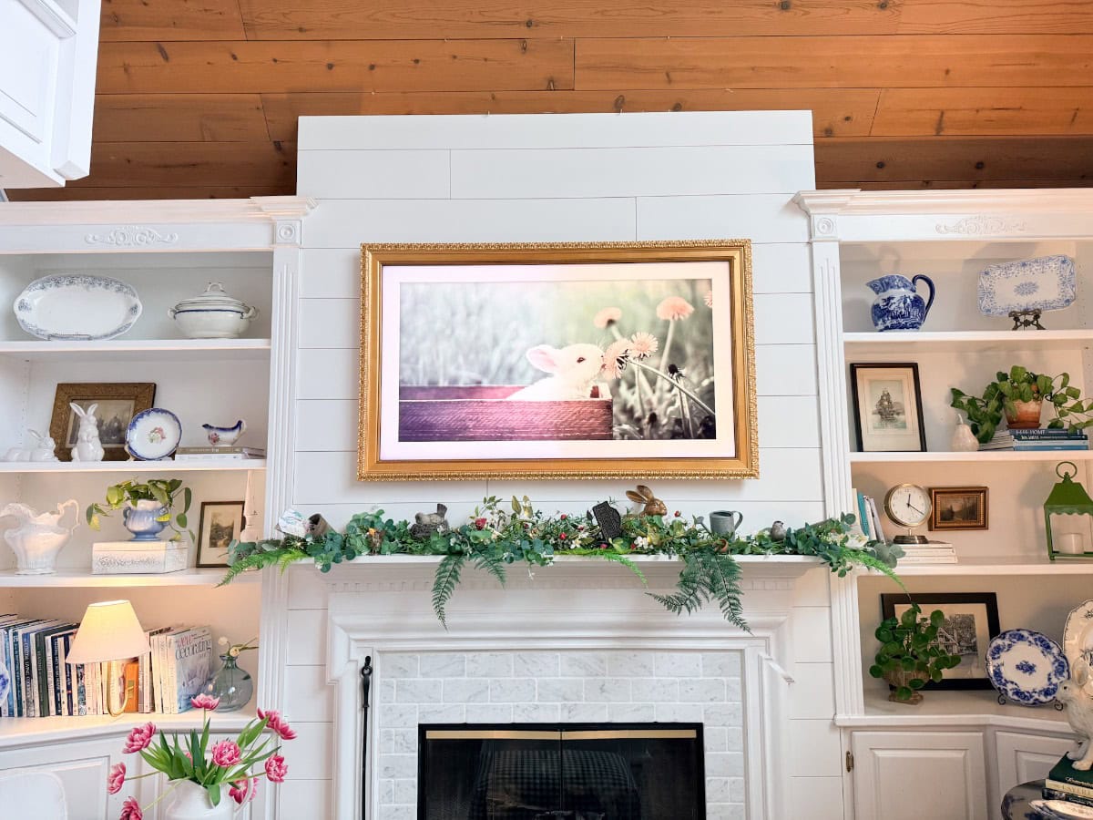 A cozy white-paneled fireplace is decorated with greenery and ferns for a charming spring home tour. Above the mantel hangs a large framed photo of a white bunny in a field, while shelves display blue and white dishes, books, and decorative items.