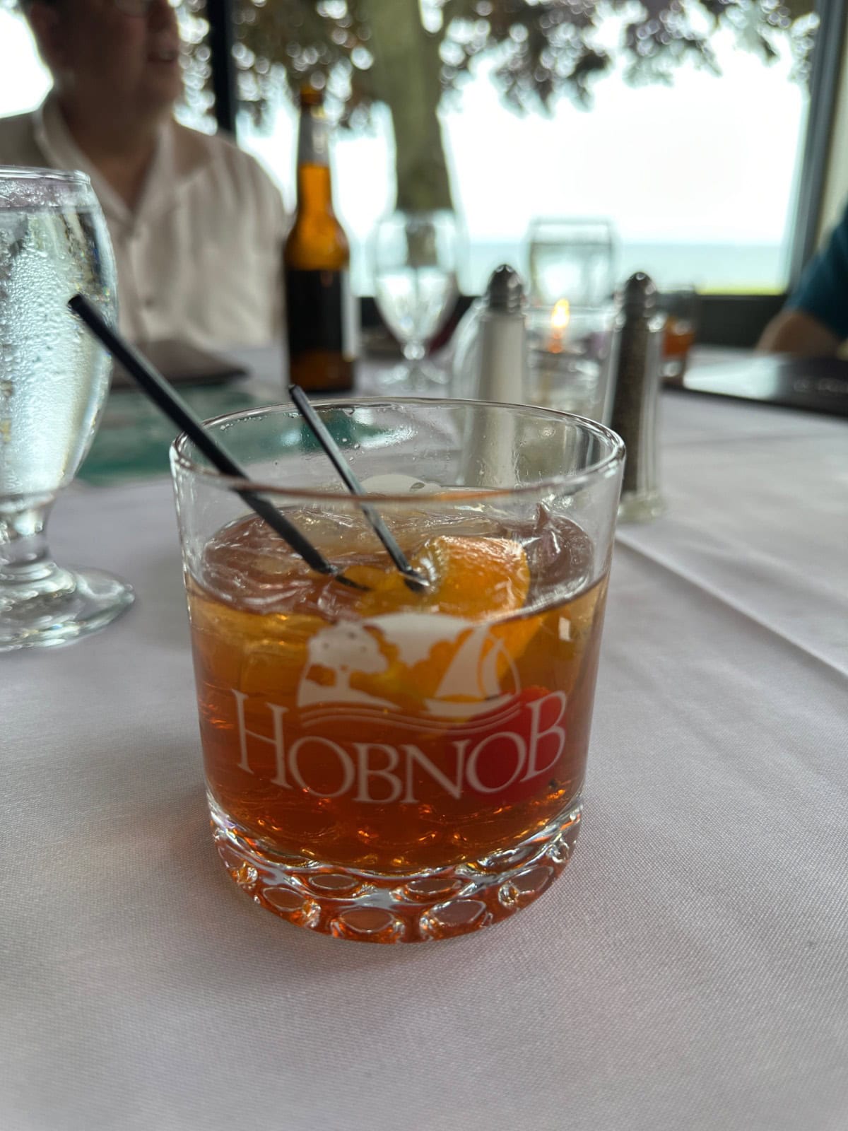 A HobNob-branded glass filled with the latest old fashioned cocktail, garnished with orange and cherries, sits on a white tablecloth at a restaurant, with a water glass and other dining items in the background.