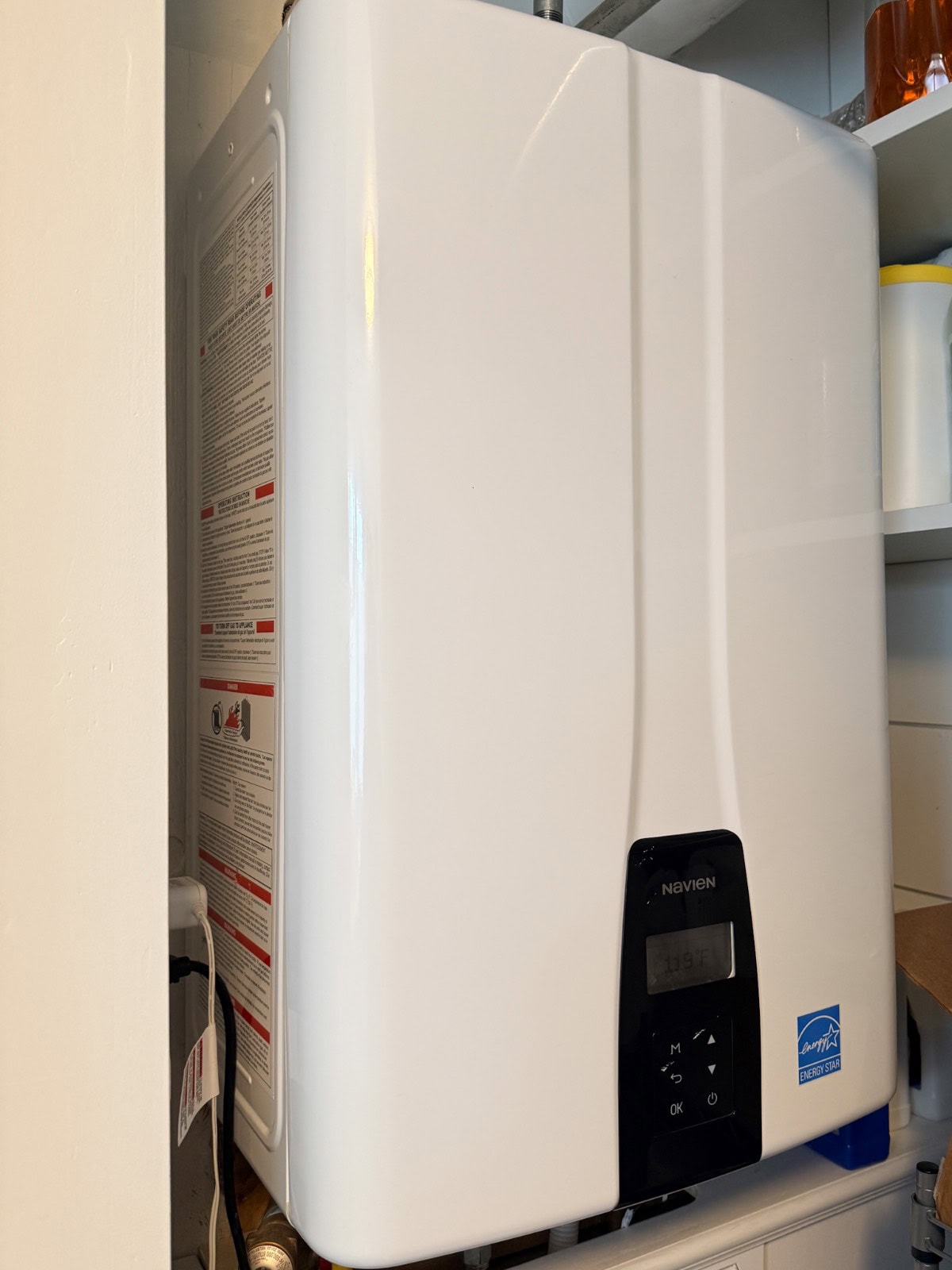 A white tankless water heater with a digital display is mounted on a wall in a small utility space, accompanied by instructions and safety information—a smart addition to any seasonal home maintenance checklist.