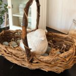 A rustic wicker basket with a curved handle holds a white ceramic bird and bluish-grey decorative eggs, creating charming vintage Easter table decor on a dark surface with a white cabinet in the background.